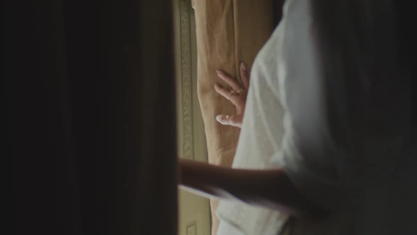Person parting brown curtain with one hand while gazing through window in soft light