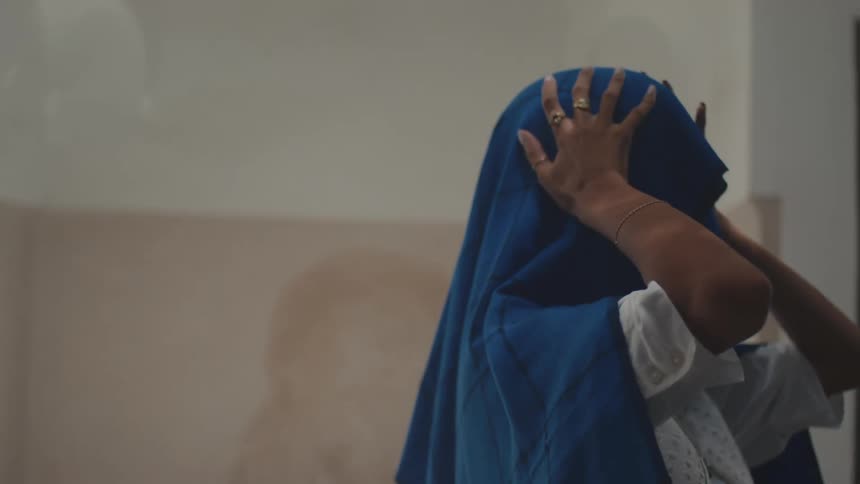 Person adjusting blue headscarf with both hands in front of light wall and green door