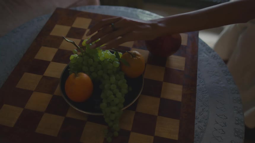 Bowl on a table with green grapes and oranges as a woman reaches over and takes grapes in her hand