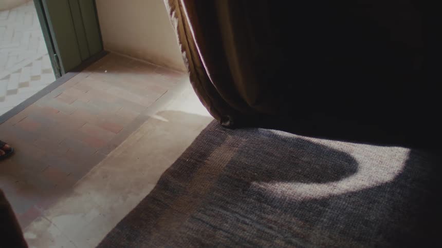 A lowangle shot shows a womans feet wearing black sandals and white clothing walking across a tile floor from a brightly lit area past a heavy dark curtain and a rug