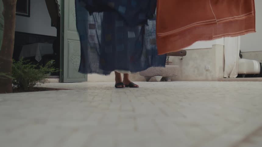 A woman in sandals and a white skirt stands on a tiled courtyard holding two pieces of hanging fabric one dark blue and one orange while the lower part of her body is visible in a lowangle shot
