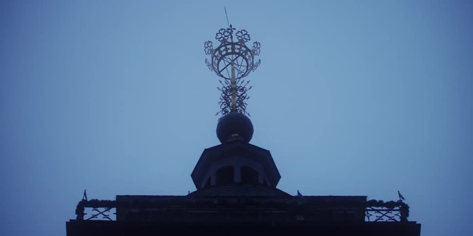 A closeup static shot features the ornate gold finial and dark cupola atop a building silhouetted against a solid overcast bluegray sky