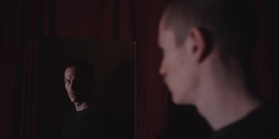 This video shows a medium shot of a young man possibly in his 20s with short reddish hair and fair skinhe is looking into a mirror in a dimly lit dramatic setting with dark red curtains or walls in the background which reflects his face with a sli