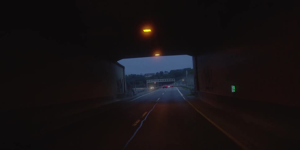 The shot is taken from a moving vehicle as it drives through a dark tunnel illuminated by overhead amber lightsthe vehicle exits the tunnel onto an open highway under a dusky or overcast sky with other traffic visible ahead