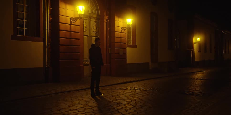 A young man standing in the street at night wearing a hoodie as light from a nearby building shines onto him