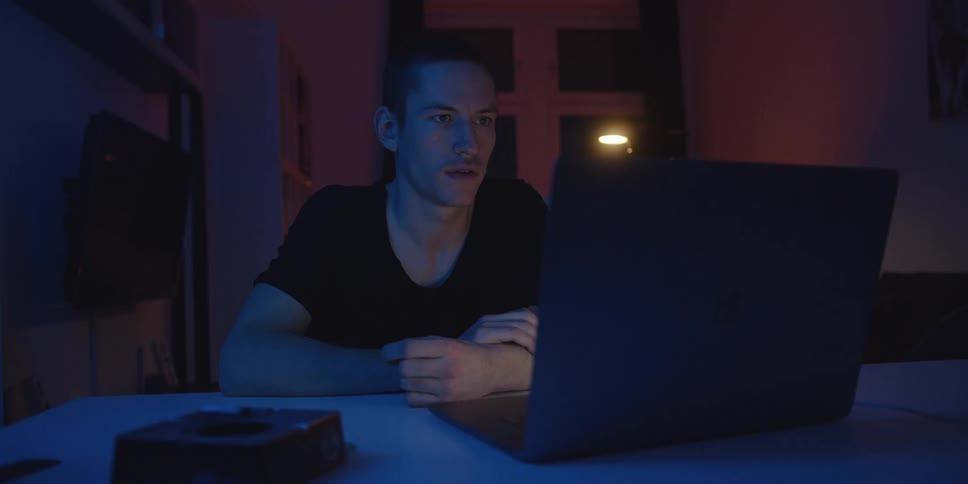 A man sits at a table focused on a laptop during a video call in a dimly lit room