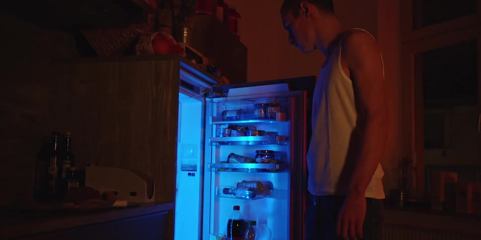 A man checks an open refrigerator at night in a dimly lit kitchen