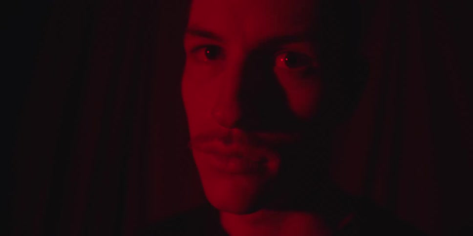 A closeup shot of a mans face bathed entirely in red light opens his eyes and stares directly ahead