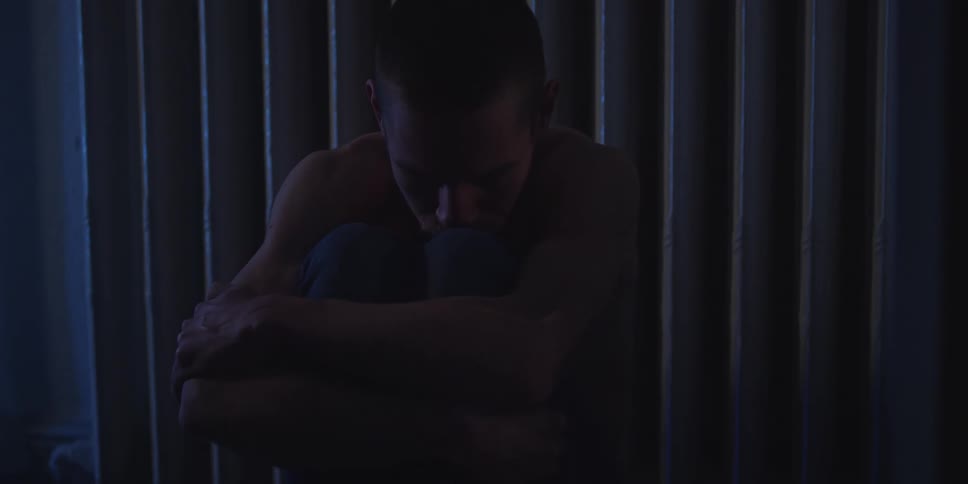 Man seated with arms wrapped around his legs in a dimly lit indoor setting