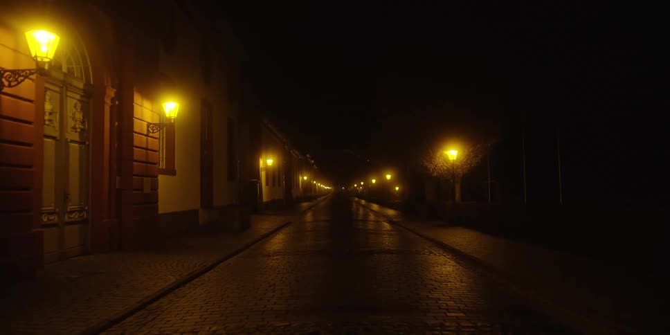 An empty wet cobblestone street lined with historic architecture and numerous bright yellow wall lanterns that cast a warm glow and reflections into the deep darkness of the night