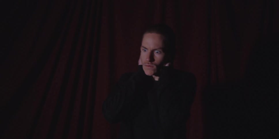 This is a closeup shot of a young man with short hair and an intense expression standing in front of dark red curtainshis expression shifts dramatically from focused intensity to apparent fear or distress as he briefly brings his hands to his head a