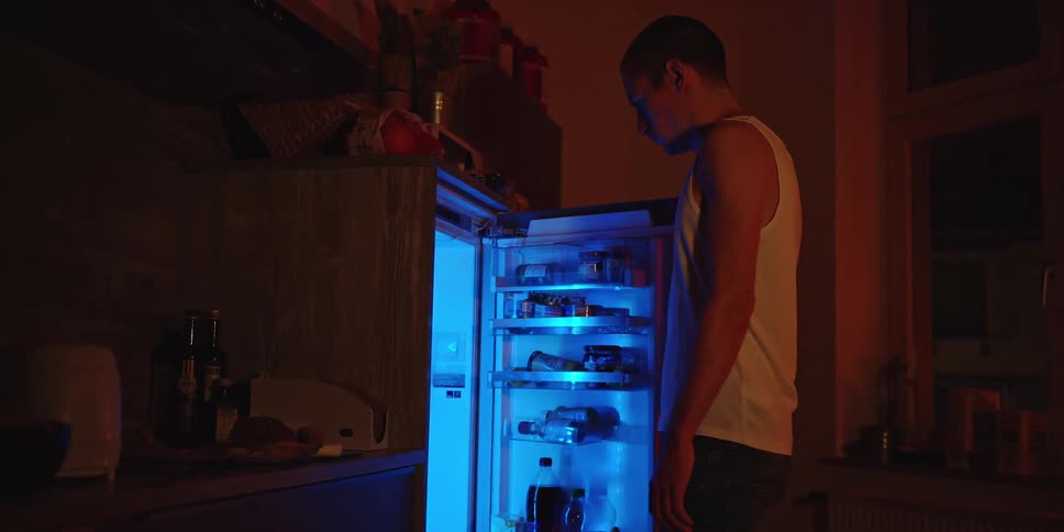A young man opening the fridge in his kitchen at night then shutting it in frustration