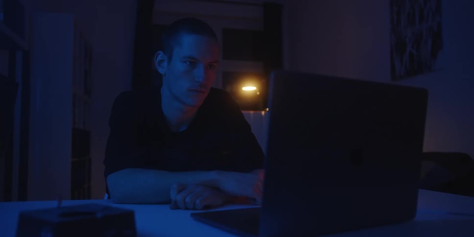 Young man sitting at a table in a dark room looking at a laptop computer