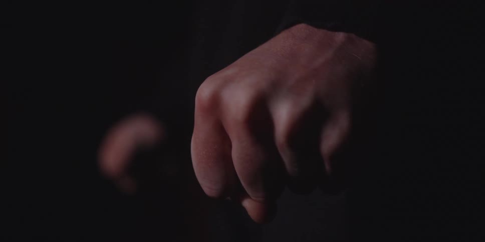 A closeup shot of a persons hand welllit from the side slowly clenches into a tight fist against a black background emphasizing the skin texture and veins