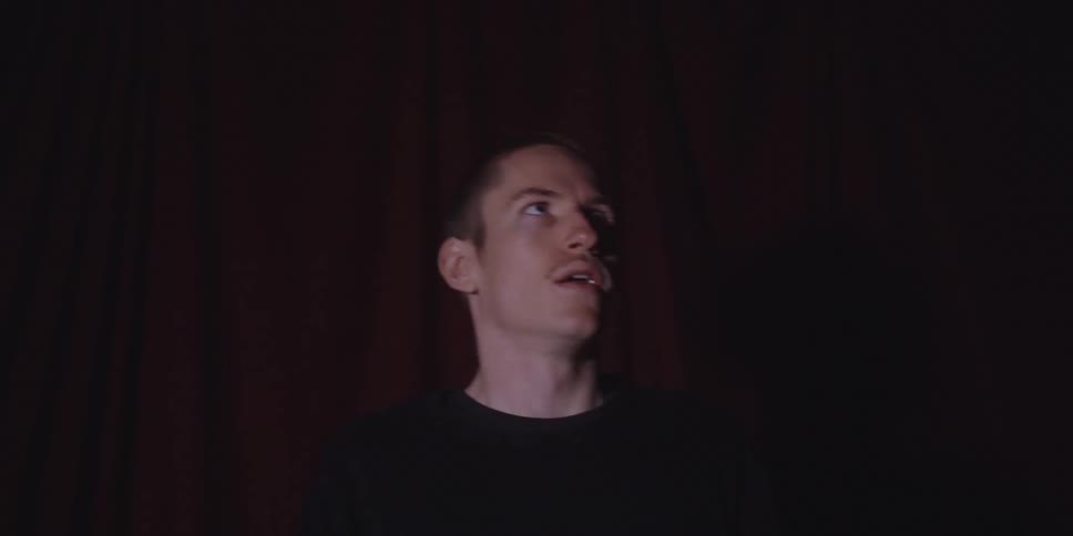 A man with a shaved head and light eyes wearing a black shirt stands in a dimly lit room against a dark red curtain looking around with intense expressive facial movements before placing his hands on his head