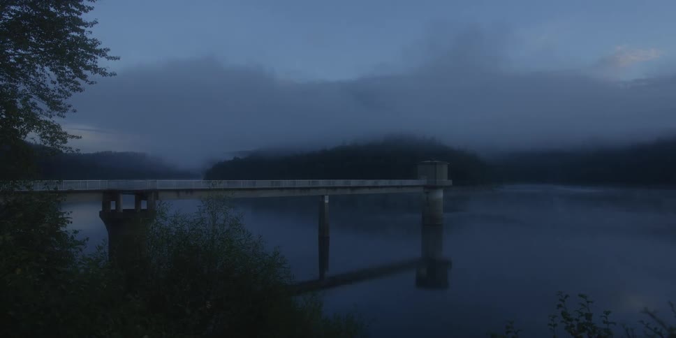 A concrete pier structure extending over a dark blue reservoir surrounded by forested hills shrouded in dense fog during dawn
