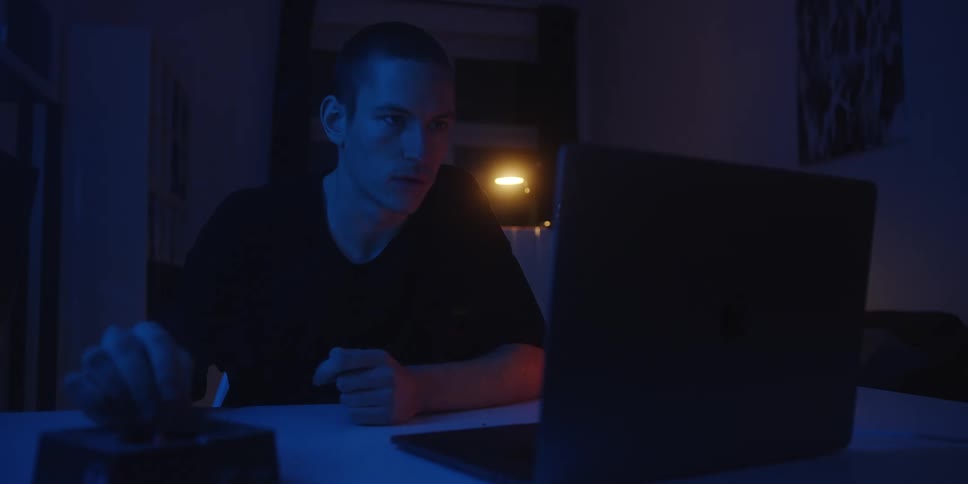 Person smoking while seated at a desk in front of a laptop indoors at night with blue lighting