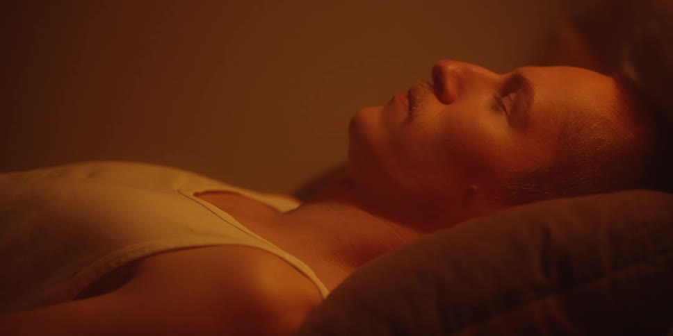 The video is a closeup shot of a person lying down seemingly asleep on a pillowthe scene is dimly lit with warm orangetoned light and the person is wearing a lightcolored tank top