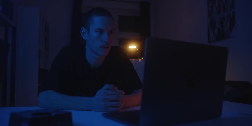 Man sits at a table watching a laptop screen in an indoor setting with blue lighting