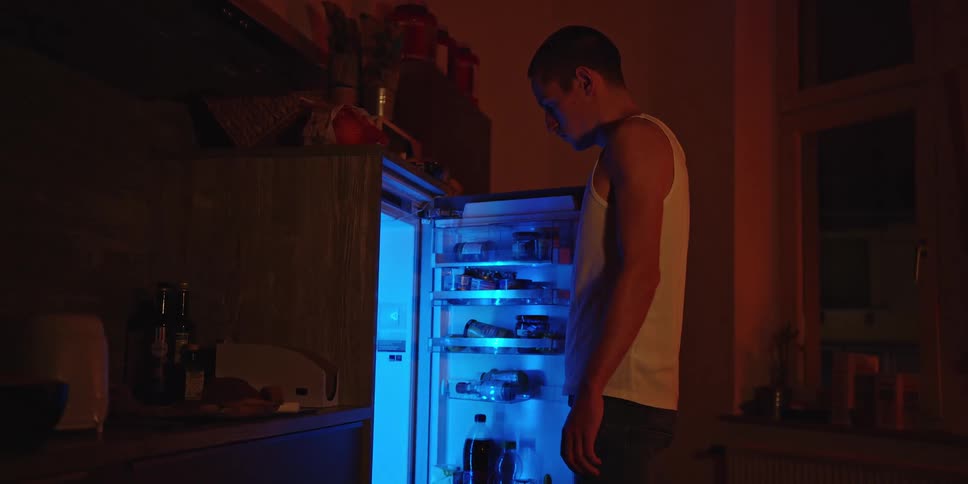 The video shows a man in a white tank top walking into a dimly lit kitchen and opening a refrigeratorthe bright blue light from the open refrigerator illuminates his face and the contents of the fridge as he looks inside