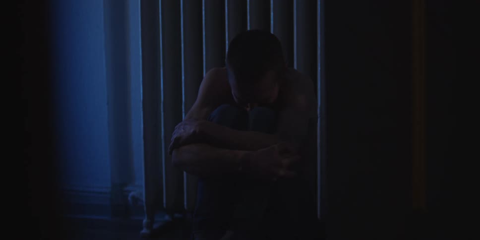Man sits on the floor hugging his knees in front of vertical blinds in an indoor setting