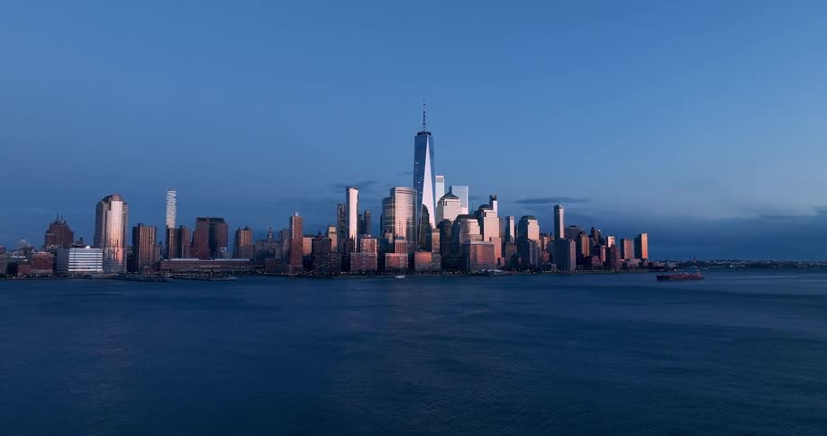 The golden light of sunset reflects brilliantly off the glass facades of the lower manhattan skyline contrasting with the deep blue hues of the twilight sky and the hudson river below