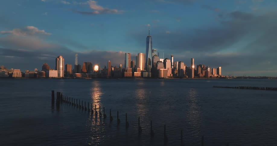 A wide sunsetlit view of a modern city skyline with a tall central skyscraper reflecting golden light dramatic clouds above and dark wooden pilings protruding from the calm blue water in the foreground