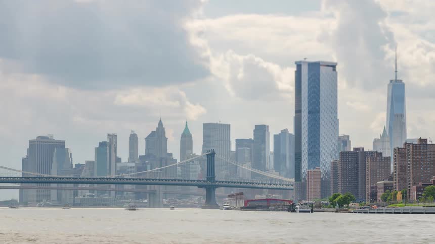 A serene timelapse captures the iconic new york city skyline and manhattan bridge as boats traverse the river and clouds drift across a cloudy sky
