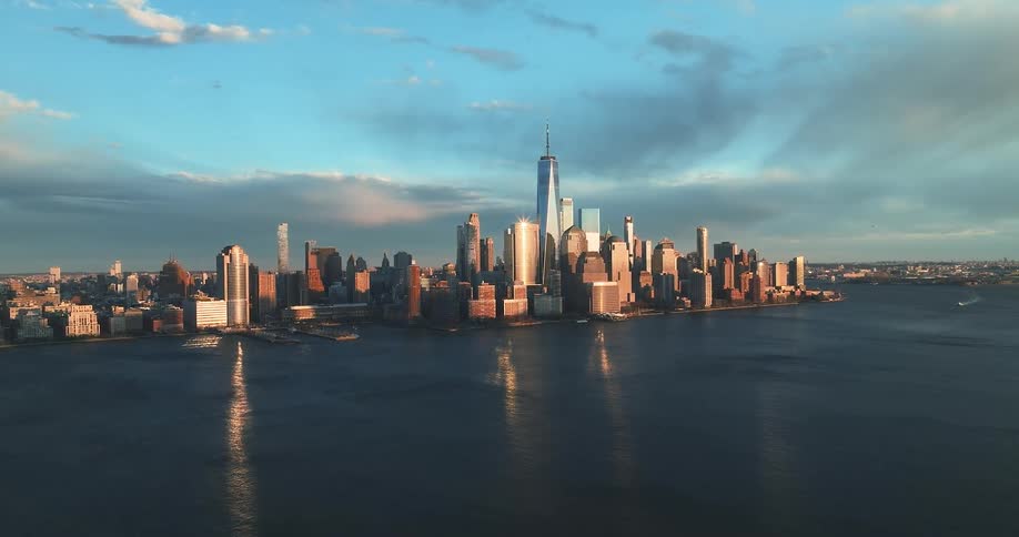 An aerial view captures the sprawling city skyline bathed in golden sunlight as a slow tracking shot moves across the calm water
