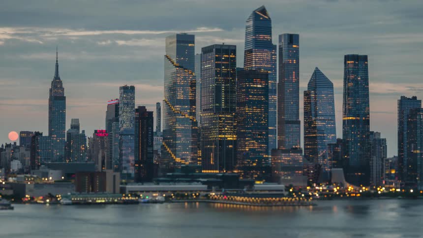A city skyline transitions from dusk to night as a large moon rises behind the buildings while the office towers begin to glow with golden interior lights against a darkening blue and orange sky