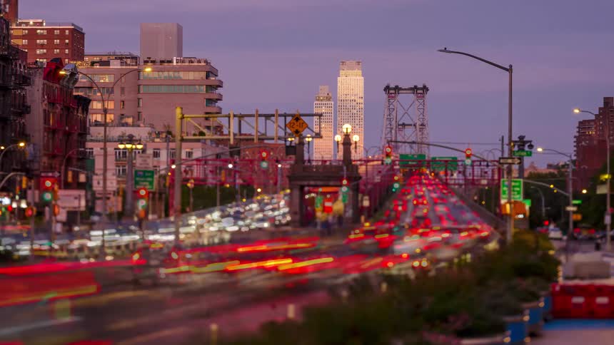 As twilight fades into night a dynamic timelapse captures the frenetic energy of new york city traffic flowing toward the williamsburg bridge transforming headlights and taillights into vibrant rivers of light against the urban skyline