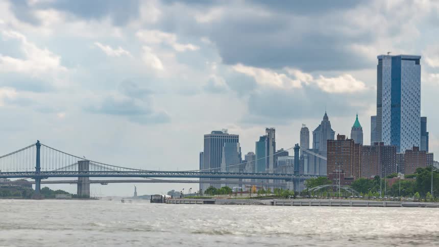 Rapidly moving storm clouds roll over the manhattan bridge and modern skyline casting shifting shadows while boats zip across the churn of the east river in this dynamic timelapse