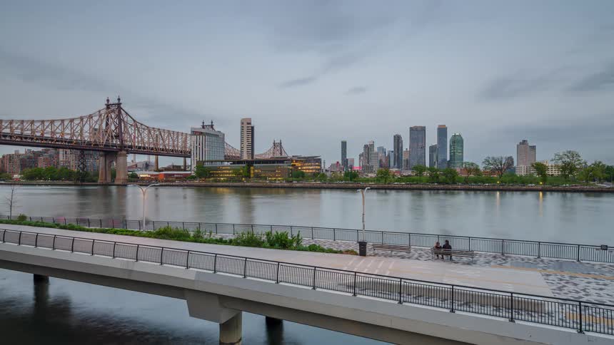 This video is a timelapse showing the transition from day to night over an urban waterfrontit features a view of a large suspension bridge the queensboro bridge59th street bridge in new york city and a skyline of highrise buildings across a river