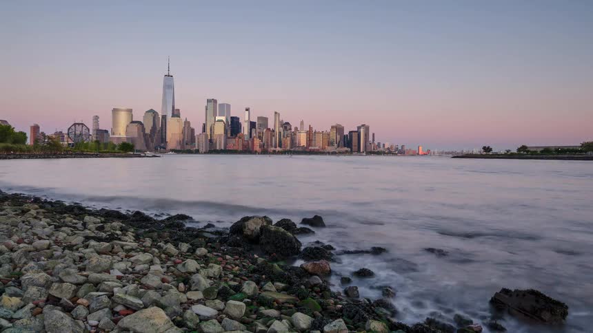 The transition from day to night over a city skyline featuring several illuminated buildings all seen from a rocky shoreline across the water