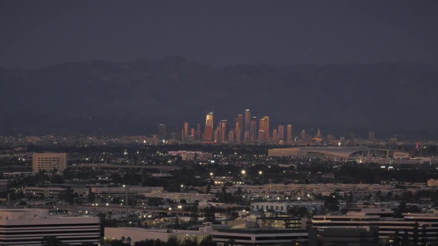 As night falls the downtown los angeles skyline glows warmly against a backdrop of dark mountains with the sprawling city lights twinkling in the vast landscape below