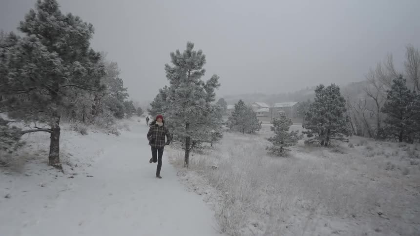 Young woman running along a snowy forest path wearing a winter jacket and hat as fresh snow falls from the sky