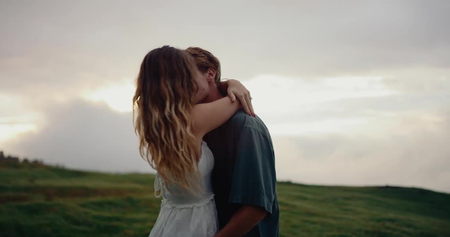 Man and a woman standing on a grassy slope on a cloudy day smile and kiss each other