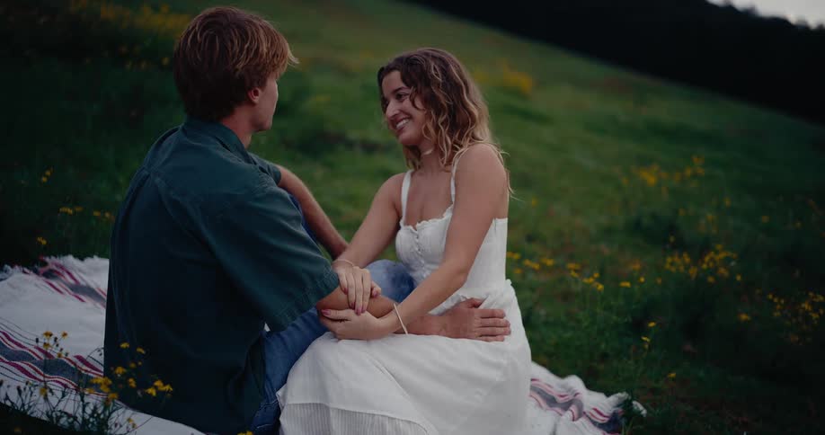 A young couple sits closely together on a striped picnic blanket in a twilight field of wildflowers sharing an intimate moment as they talk and gaze affectionately at one another