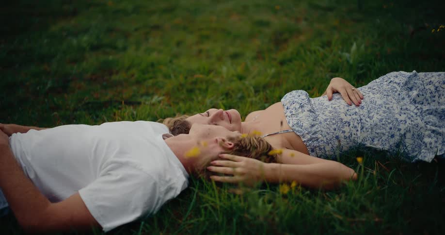 A young couple lying on a grassland positioned opposite each other with their heads side by side as the woman gently runs her hand through the mans hair