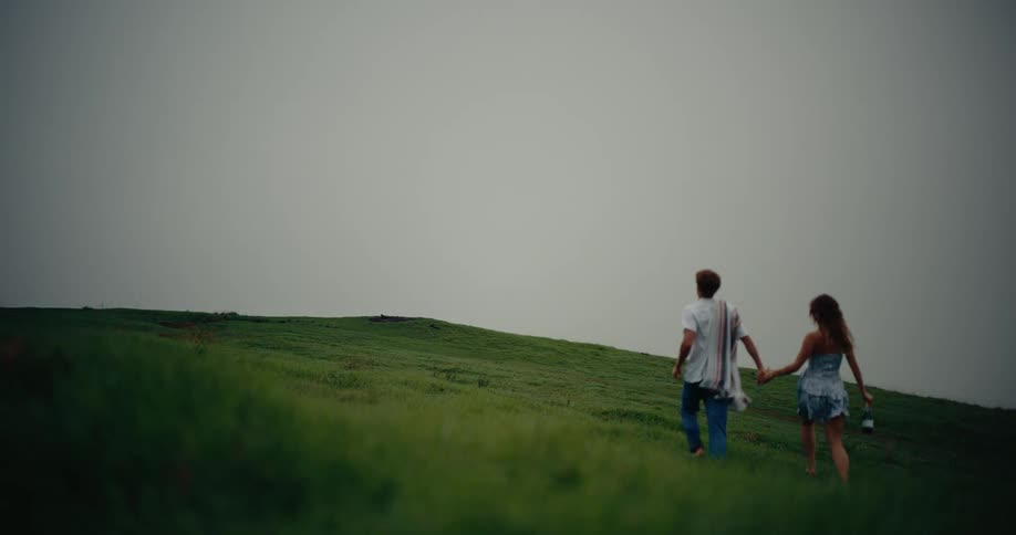 A couple runs hand in hand up a grassy hill toward a foggy horizon under an overcast sky
