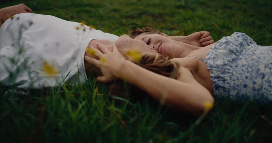 A young man and woman lie together in a grassy field of wildflowers smiling and looking into each others eyes while she gently strokes his face during an intimate quiet moment outdoors
