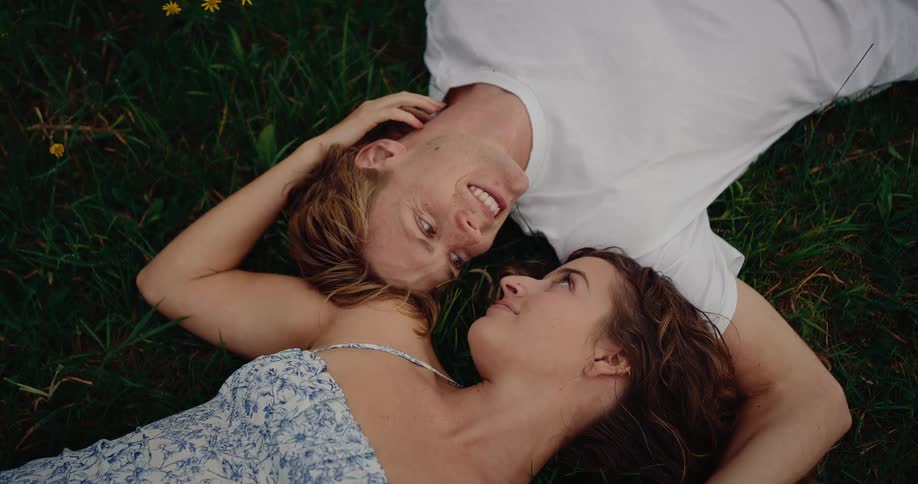 A couple lies in a lush green field dotted with small yellow flowers sharing a passionate gaze as the woman gently caresses the back of the mans head while he smiles warmly at her