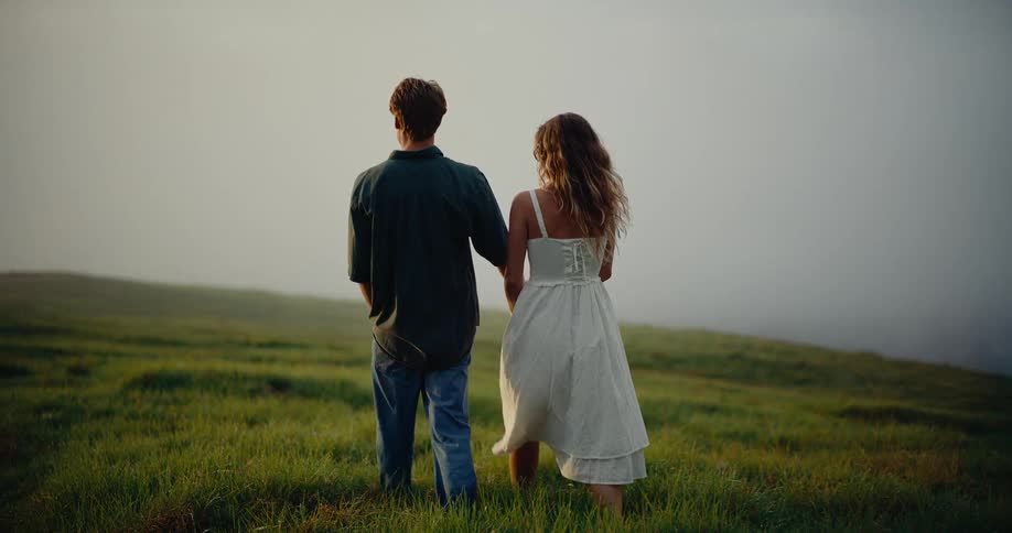 A young couple walking handinhand across a grassland at sunset warm light stretching softly around them