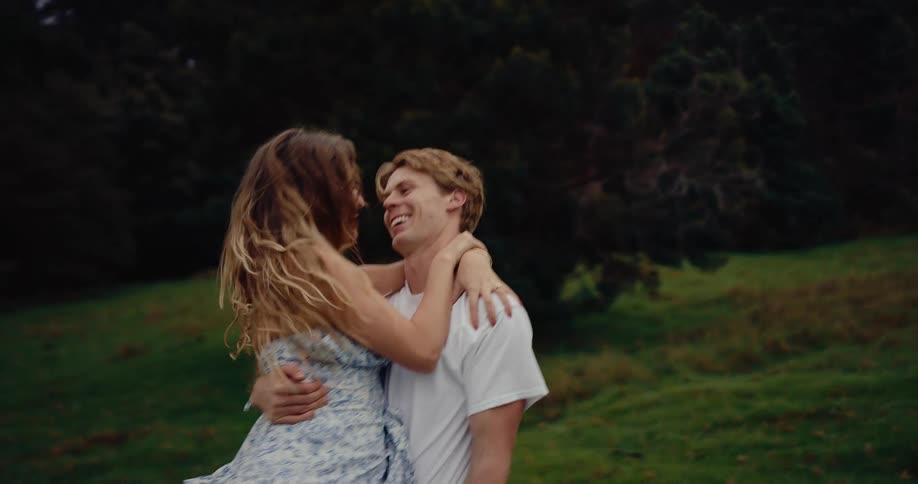 A young couple joyfully runs and embraces in a grassy treed area under an overcast sky