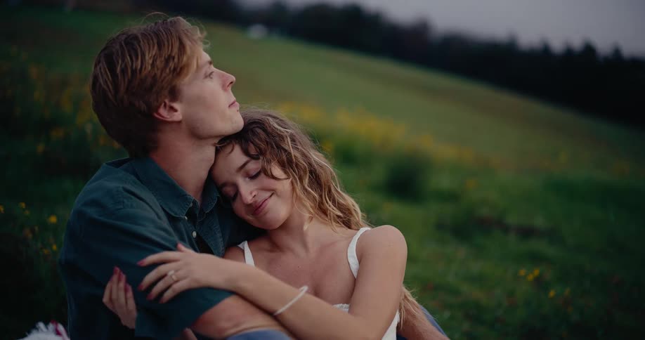 A loving couple embraces in a wildflowerdotted field at dusk sharing a gentle forehead kiss as they gaze into the distance together