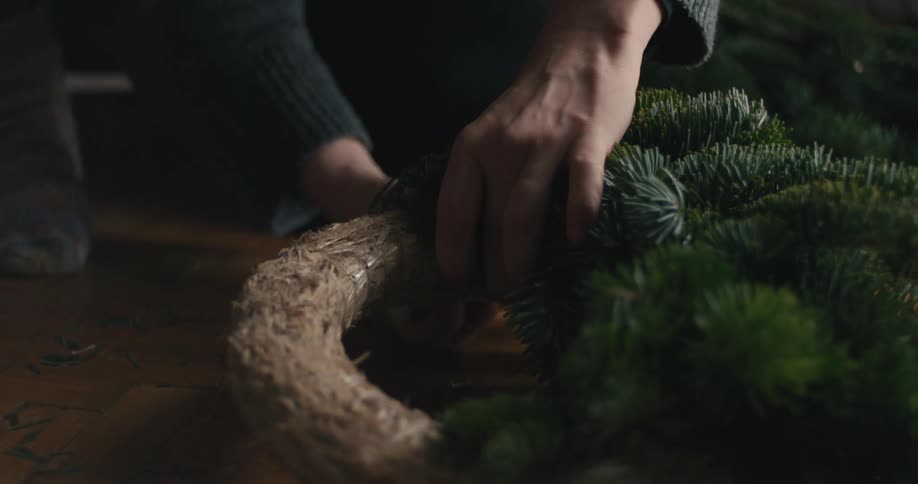 A woman wearing a grey sweater sits on a wooden floor and uses her hands to carefully attach fresh green pine branches onto a circular straw base to create a decorative holiday wreath
