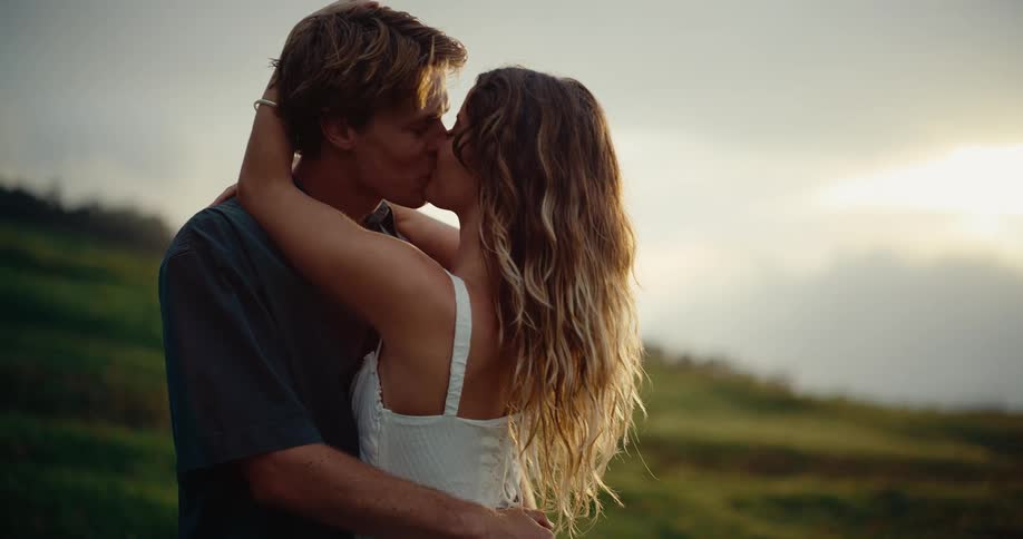 A young couple embraces and shares a kiss outdoors against a sunset or sunrise backdrop over a hilly landscape