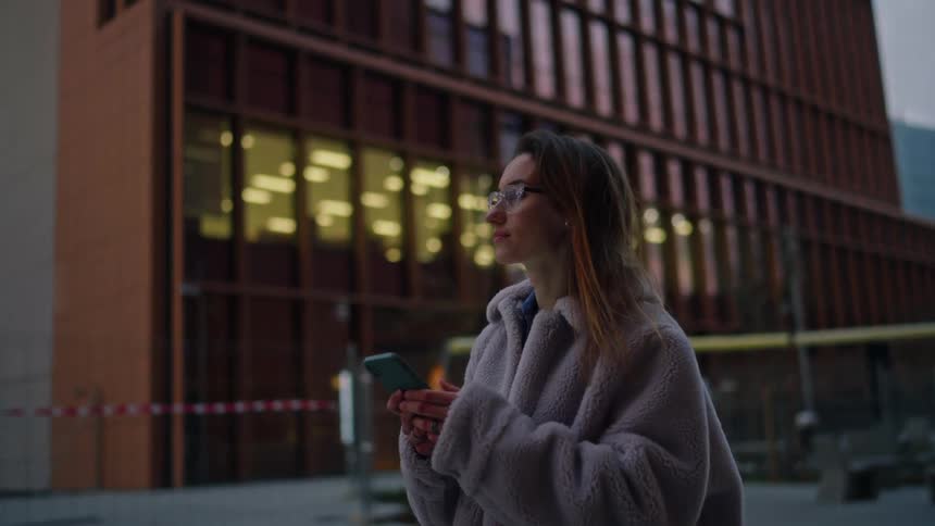 As dusk settles a woman checks her smartphone while walking through a modern urban plaza the warm glow of an office building reflecting in her glasses