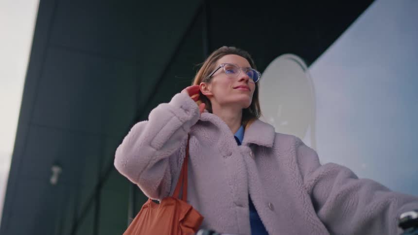 A woman with glasses and a lilac fleece coat walks with a bicycle past a modern glass building on a cloudy day