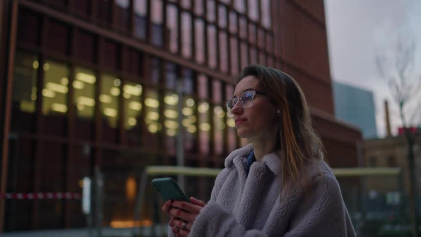 A woman wearing glasses and a fuzzy coat looks at her phone while standing outdoors in an urban area with modern and older buildings around